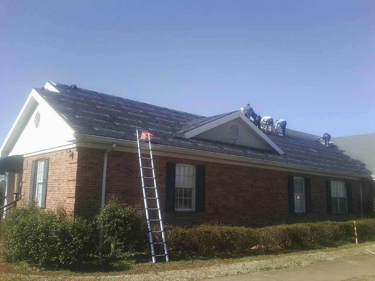Crew tearing shingles off a brick ranch roof with a ladder set — Metal Roof Repair work by our crew in Boswell